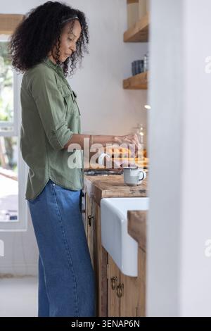 Afroamerikanerin in grünem Hemd, die Getränk in Becher auf der Theke in der Bauernküche rührt Stockfoto