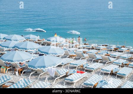 Nizza Frankreich, 31 2026, mit blau-weiß gestreiften Sonnenliegen und passenden Sonnenschirmen am Kiesstrand von Nizza, Frankreich, mit Leuten, die im Wasser schwimmen Stockfoto