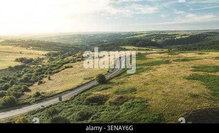 Eine einsame Asphaltstraße, die sich durch eine weite, hügelige Landschaft mit üppigen grünen Feldern und weit entfernten Wäldern, die in sanfte S getaucht sind, führt aus der Vogelperspektive Stockfoto