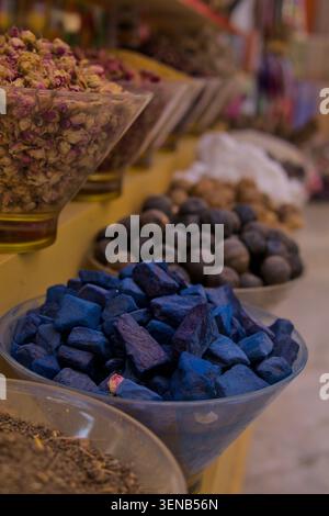 Lebendige Gewürze und getrocknete Blumen füllen Glasschalen. Blaue Würfel stehen im Kontrast zu Erdnüssen und Samen. Warme Beleuchtung hebt Texturen und Farben hervor. Stockfoto