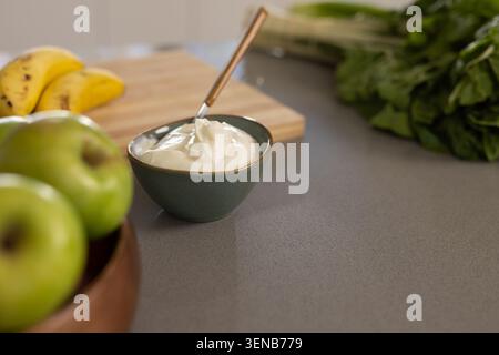 Kleine grüne Schüssel mit dicker Creme, Löffel auf grauer Theke, grüne Äpfel, Bananen verschwimmen Stockfoto