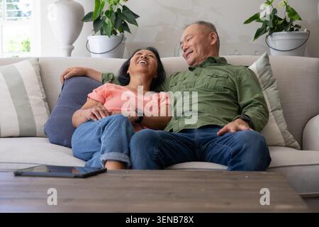 Asiatisches Paar, reifer Mann und ältere Frau, sitzen zu Hause auf hellem Sofa und lachen in der Nähe von Tablets Stockfoto
