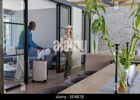 Ein Paar, das durch schwarze Glastüren ins Loft geht, eine Frau mit Hut, die zurückblickt, ein Mann, der den Koffer drückt Stockfoto
