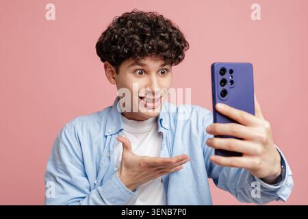 Ein fröhlicher junger Mann mit lockigen Haaren interagiert aufgeregt mit seinem Smartphone und zeigt den modernen Trend von Videoanrufen und Social-Media-Interaktionen Stockfoto