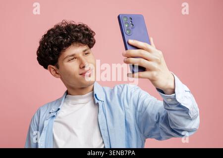 Ein junger Mann mit lockigen Haaren lächelt, während er mit seinem Smartphone ein Selfie macht und einen verspielten und modernen Lebensstil vor einem leuchtend rosa Backdro zeigt Stockfoto
