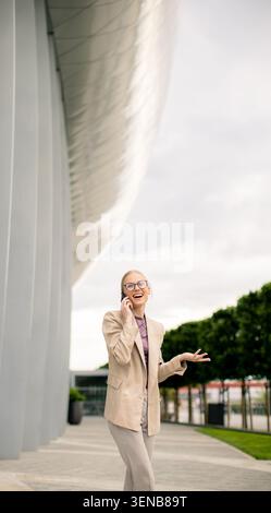 Eine Geschäftsfrau steht vor einem hohen Gebäude mit einem geschwungenen Dach. Sie redet auf ihrem Handy und lächelt, während sie eine Brille trägt. Grüne Bäume säumen das Gebiet, Stockfoto