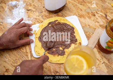 Die Hände der Person bereiten einen süßen Pfannkuchen zu, verteilen reichhaltige Schokoladen-Haselnusscreme auf der goldenen Oberfläche, mit einem erfrischenden Zitronengetränk und Marmeladenglas vor Stockfoto