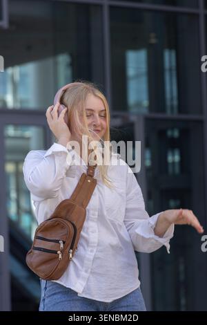 Eine Erwachsene Frau, die vor einem Glasgebäude steht, trägt rosa Kopfhörer und hält eine braune Tragetasche Stockfoto
