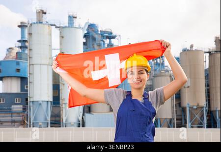 Junge Ingenieurin im Helm, die Staatsflagge der Schweiz schwenkt, während sie vor großen Tanks in der Chemiefabrik steht Stockfoto