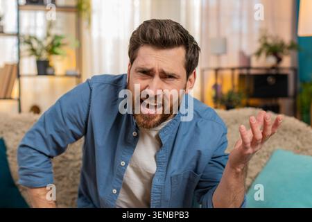Ein junger Mann sitzt zu Hause auf dem Sofa, schwinzt mit Verdacht in die Kamera und zweifelt, was er sieht. Der Kaukasier sieht skeptisch aus, verengt die Augen ungläubig und unsicher wegen unerwarteter Informationen Stockfoto