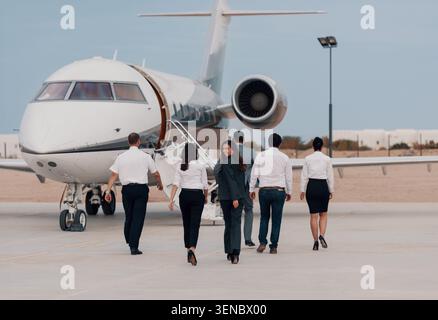 Das Business-Team und die Flugbesatzung laufen in der Abenddämmerung auf einem Flughafenparkett zu einem Privatjet, wobei eine Frau zurückblickt und lächelt, ideal für Geschäftsreisen Stockfoto