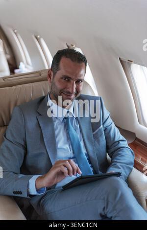 Lächelnder Geschäftsmann aus dem Nahen Osten in grauem Anzug mit einem digitalen Tablet an Bord einer luxuriösen Privatjet-Kabine mit weicher Fensterbeleuchtung, ideal für Geschäftsleute Stockfoto