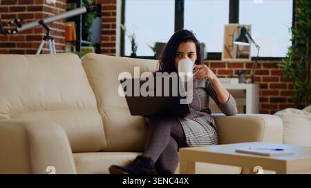 Telearbeiter, der auf der Couch liegt und Kaffee trinkt, während er Aufgaben für das Online-Lernen aus der Ferne erledigt. Stockfoto