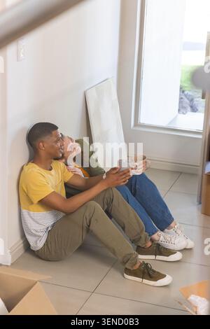 Verschiedene Pärchen sitzen auf gefliestem Boden neben dem Fenster und halten Tassen zwischen Umzugskartons und angehobener Leinwand Stockfoto