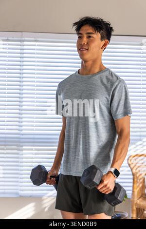 Asiatischer Mann mit athletischem T-Stück, der Hexenhülsen hebt, die im Wohnzimmer bei Jalousien mit Uhr stehen Stockfoto