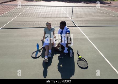 Diverse Tennispartner sitzen auf dem Platz am Netz in Tennisbekleidung mit Schlägern und Wasserflasche Stockfoto