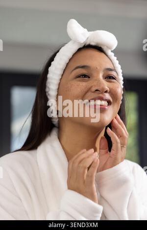 Asiatische Frau mit Plüschkopfband und Gewand, das Gesicht mit goldenem Ring am Eitelbereich in der Nähe des Fensters berührt Stockfoto