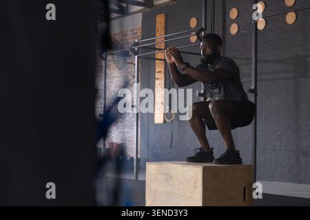 Afroamerikanischer Mann hockt auf hölzernen Plyoschachteln im Fitnessstudio, trägt ein schwarzes Sporthemd, Kopierraum Stockfoto