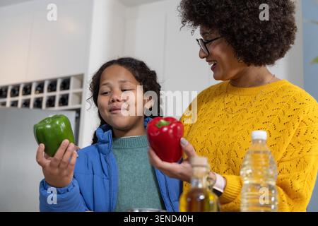 Verschiedene Mutter und Tochter halten grüne und rote Paprika in der Nähe der Ölflasche auf der Theke und untersuchen sie Stockfoto