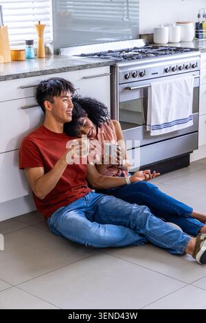 Verschiedene Paare sitzen auf gefliestem Boden und lehnen sich an Schränke in der Küche und halten Keramikbecher Stockfoto