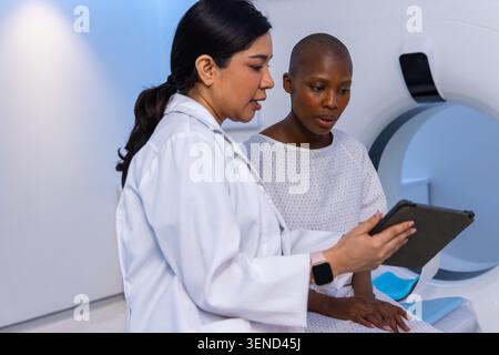 Ärztin mit Laborkitt, der weibliche Patientin im Kleid am Scanner in der Radiologie eine Tablette zeigt Stockfoto