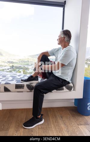 Senior Mann sitzt auf gemusterter Fensterbank und blickt auf das Tal mit blauer Schaumwalze, Kopierraum Stockfoto