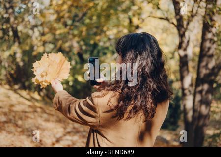 Die Person nimmt ein Smartphone-Foto von gelben Herbstblättern auf, während sie im Park lächelt. Social Media Influencer Lifestyle, Herbst Bloginhalte, Lifestyle Stockfoto