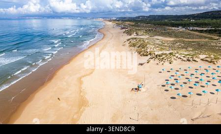 Costa Caparica, Lissabon. Blick aus der Vogelperspektive auf den Strand Costa Caparica, Portugal. Goldene Sandkurve entlang des Atlantischen Ozeans. Blaue Regenschirme an der Küste. Stockfoto
