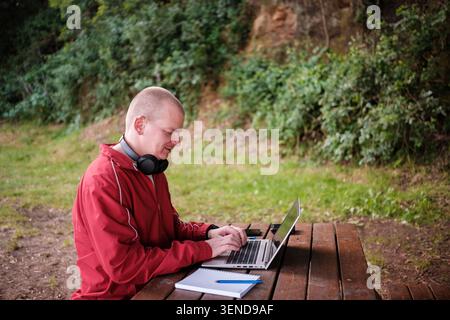 Mann tippt auf einem Laptop mit Kopfhörern an einem hölzernen Picknicktisch draußen Stockfoto