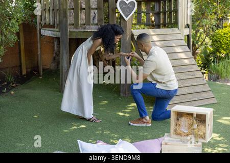 Ein Paar kniet im Garten, ein Mann präsentiert eine offene Ringbox, eine Frau mit weißem Kleid am Spielset Stockfoto