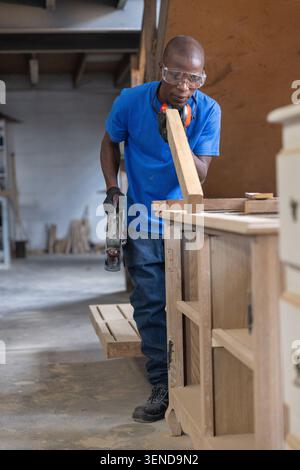 Afroamerikanischer Zimmermann mit Hobelmaschine auf Holzdiele mit Sicherheitsausrüstung im Geschäft mit Werkbank Stockfoto