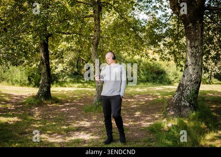 Mann, der im Freien in einem Park steht und ein Hörbuch oder Musik mit kabellosen Kopfhörern hört Stockfoto