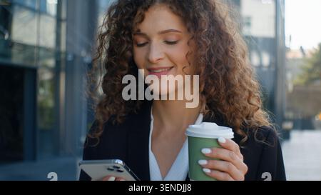 Lächelnde Gadget-süchtige kaukasische Geschäftsfrau, die im Handy sucht, Kaffee trinkt, morgens zur Arbeit geht in der Stadt Geschäftsfrau weibliches Mädchen Stockfoto