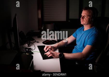 Mann mit Kopfhörern und blauem T-Shirt, der in einem dunklen Raum auf der Tastatur schreibt Stockfoto