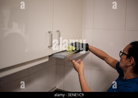 Frau in Handschuhen, die eine Dunstabzugshaube und Küchenschränke mit einem Tuch reinigen Stockfoto