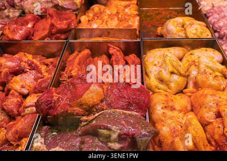 Frisch marinierte rohe Fleischprodukte, einschließlich Hühnerflügel, Schweinerippchen und Würstchen in Metallbehältern. Auswahl an gewürzten Fleischsorten in einem Supermarkt Stockfoto