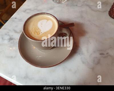 Schaumkaffee mit herzförmiger Latte Art, serviert in einer Keramikschale und Untertasse auf einem weißen Marmortisch, was an gemütliche Café-Atmosphäre und Mornin erinnert Stockfoto