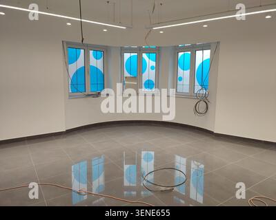 Innenansicht eines leeren Geschäftsbereichs während der Renovierung, mit glänzend grauen Bodenfliesen mit hellen Fensterreflexen, geschwungenen weißen Wänden, Stockfoto