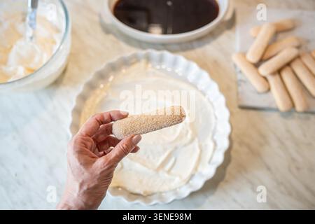 Blick von oben auf die Frauenhand, die Ladyfinger Kekse über cremigem Boden hält, während hausgemachtes Tiramisu auf Marmoroberfläche zubereitet wird. Dessertzubereitungsprozess mit Kopierraum für Rezept- und Lebensmitteldesign Stockfoto