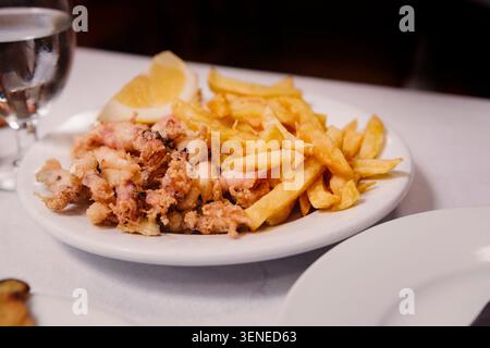 Knusprig gebratener Baby-Tintenfisch und Pommes frites serviert mit Zitronenkeil auf weißem Teller im spanischen Restaurant. Mediterrane Meeresfrüchte. Stockfoto