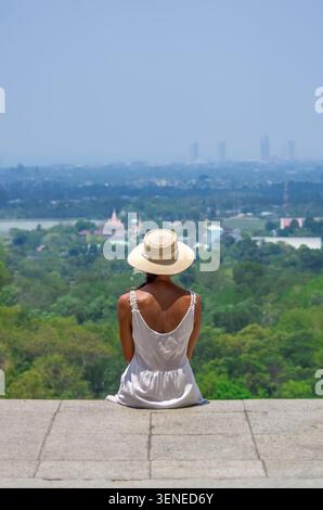 Eine junge Frau in weißem Kleid und Strohhut sitzt auf einem Steinberg mit Blick auf eine riesige tropische Stadtlandschaft mit üppigem Grün, Tempeln und einem Stockfoto