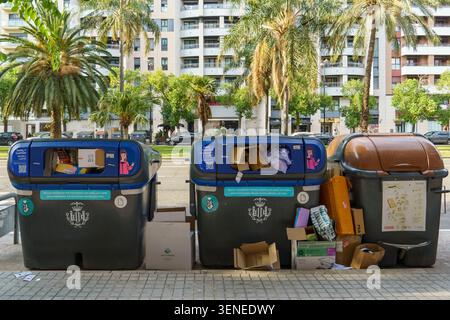 Große Mülltonnen gefüllt mit Pappe auf einer Stadtstraße in Valencia, Spanien, mit Palmen und modernen Gebäuden im Hintergrund Stockfoto