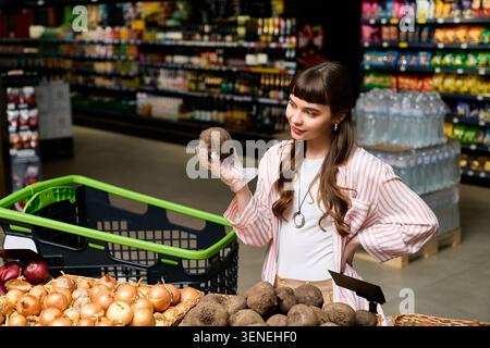 Junge attraktive Frau in stilvollem Outfit untersucht fröhlich frisches Gemüse im Laden. Stockfoto