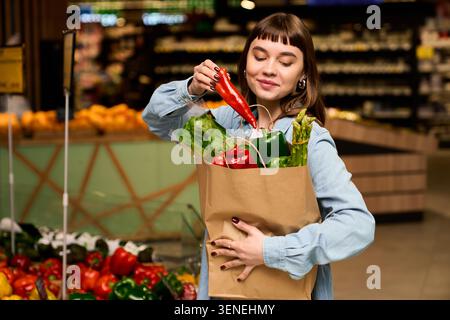 Die freudige Frau pflückt gerne frisches Gemüse in einem geschäftigen Supermarkt voller bunter Produkte. Stockfoto