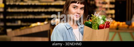 Eine junge Frau mit schickem Stil hält beim Einkaufen eifrig eine Tasche voller buntem Gemüse. Stockfoto
