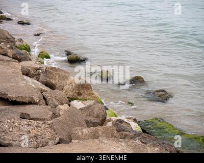 Eine detaillierte Nahaufnahme der felsigen Küste, wo verwitterter Beton und Naturstein auf das ruhige Meer treffen. Große Felsen sind teilweise mit V bedeckt Stockfoto