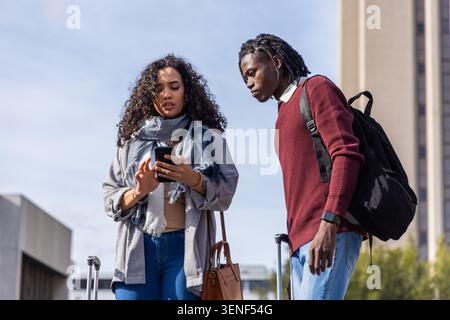 Verschiedene Freunde stehen auf dem plaza, eine Frau, die das Telefon hält, während sich der Mann lehnt, und zeigt Koffer Stockfoto