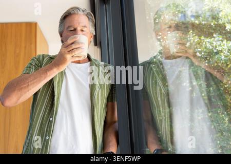 Reifer erwachsener Mann, der im Haus in der Nähe einer Glastür steht und aus dem Becher schlürft und ein grünes gestreiftes Hemd trägt Stockfoto