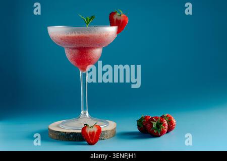 Frische Erdbeerdaiquiri in einem Glas auf einem Holzständer mit ganzen Beeren auf blauem Hintergrund Stockfoto