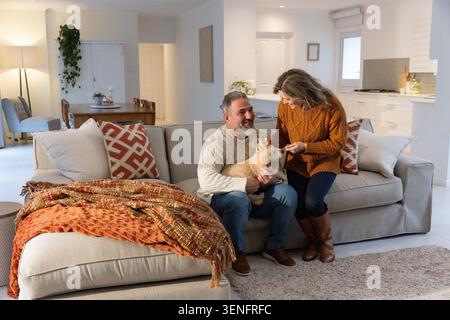 Ein Paar sitzt zu Hause auf einem hellgrauen Schnittsofa, eine Frau, die sich zu einem kleinen, braunen Hund geneigt hat Stockfoto